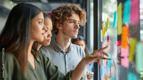 professionals in a brainstorming session, with a focus on a young man with curly hair pointing at sticky notes on a glass wall, indicating a collaborative work environment