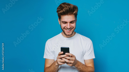 young man with styled hair, smiling while looking at his smartphone against a solid blue background © MP Studio
