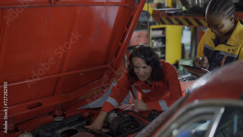 Two women are working on a red car engine. One of them is smiling. The other woman is looking at her phone
