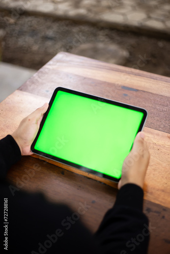 Mock up photo of a close up shot featuring a mans hand holding an iPad tablet with a green screen against the background of a wood cafe table
