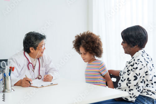 Pediatrician with glasses smiles at a young child, with caring mother by their side in bright clinic. Cheerful doctor discussing health with child and her attentive mother in well-lit medical office