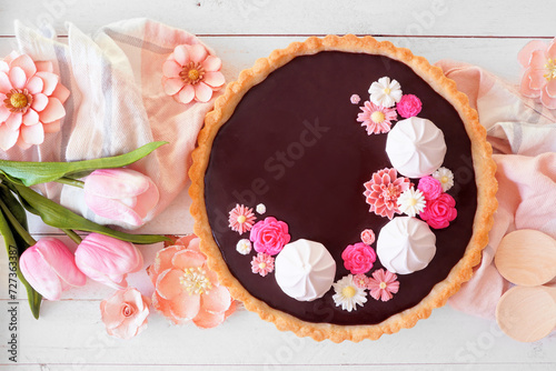 Gourmet chocolate tart with meringue and pink chocolate flowers. Overhead view table scene on a white wood background. Spring baking concept.