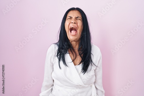 Mature hispanic woman standing over pink background angry and mad screaming frustrated and furious, shouting with anger. rage and aggressive concept. © Krakenimages.com