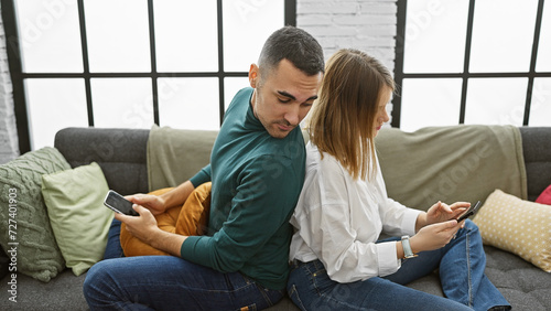 A couple sits closely on a couch, with the man peeking at the woman's phone in a modern living room setting.
