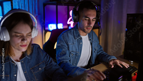 A man and woman focused on gaming with headphones in a dark room illuminated by colorful neon lights.