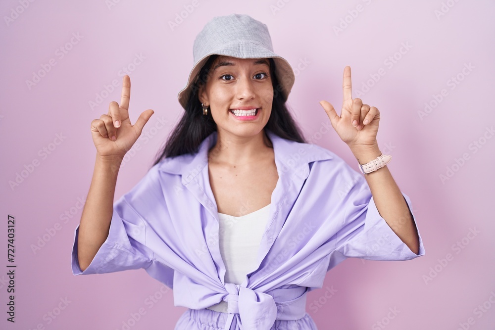 Young hispanic woman standing over pink background wearing hat smiling amazed and surprised and pointing up with fingers and raised arms. Young hispanic woman standing over pink background wearing hat smiling amazed and surprised and pointing up with fingers and raised arms.