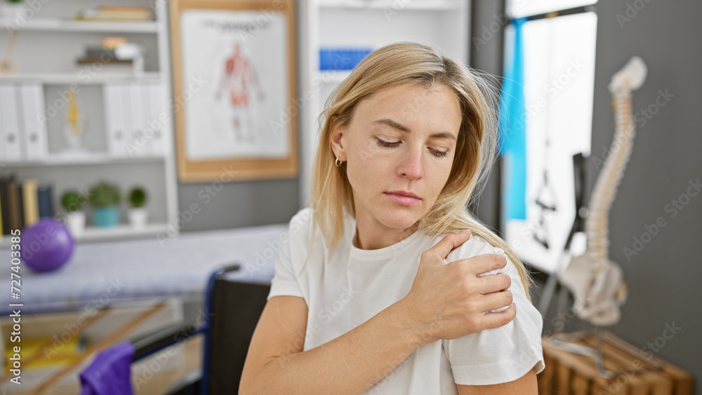 A young blonde woman showing discomfort in her shoulder at a modern physiotherapy clinic A young blonde woman showing discomfort in her shoulder at a modern physiotherapy clinic