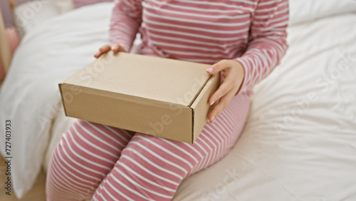 A young woman in striped pajamas holds a cardboard package while sitting on a bed with a white duvet in a cozy bedroom setting.