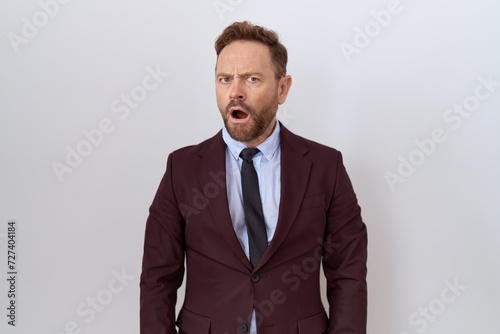 Middle age business man with beard wearing suit and tie in shock face, looking skeptical and sarcastic, surprised with open mouth