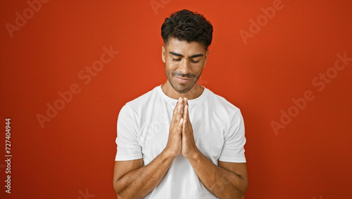 A young hispanic man with a beard in a white t-shirt against a solid red background expresses gratitude with a namaste gesture. photo