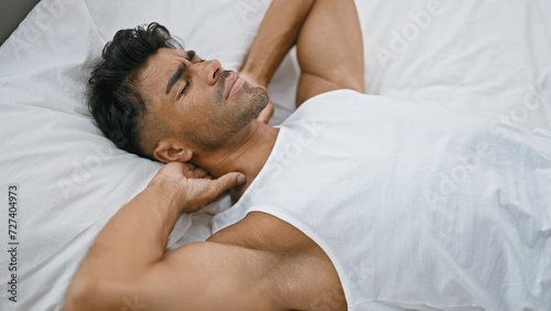 A young hispanic man with a beard sleeps soundly in a white bedroom, depicting rest, comfort, and indoor tranquility.