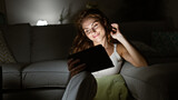 A smiling woman enjoys tablet entertainment comfortably on her living room couch at night