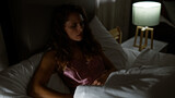 A thoughtful young woman sits in bed with a lamp on in a dimly lit bedroom, imparting a contemplative mood.