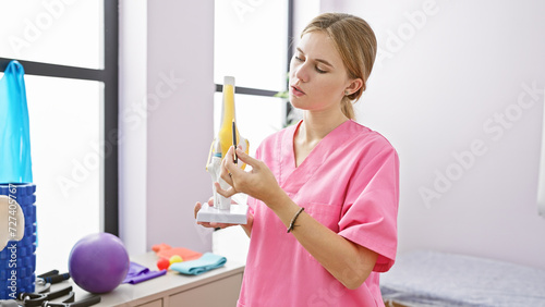 A blonde woman in pink scrubs examines medical equipment in a bright rehabilitation clinic room
