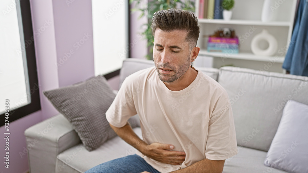 Worried young hispanic man suffering stomachache, touching aching belly while sitting on living room sofa. unhappy adult indoors, serious illness worry at home. Worried young hispanic man suffering stomachache, touching aching belly while sitting on living room sofa. unhappy adult indoors, serious illness worry at home.