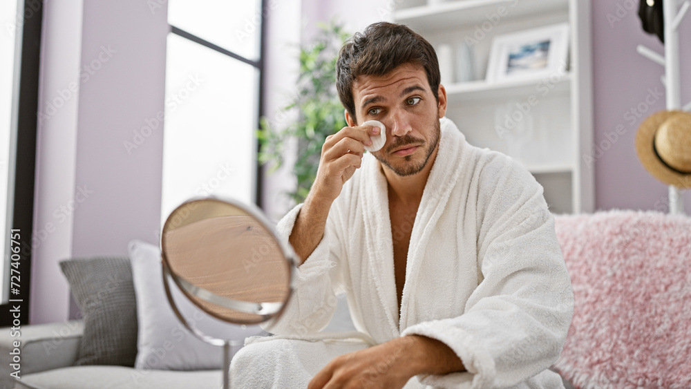 Handsome hispanic man in a white bathrobe applying skincare in a well-lit modern apartment. Handsome hispanic man in a white bathrobe applying skincare in a well-lit modern apartment.