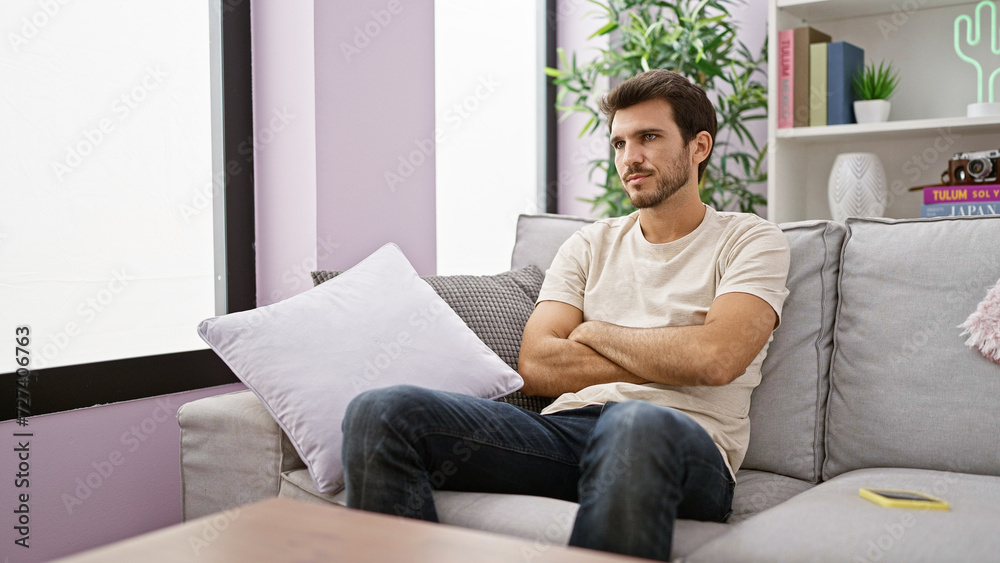 A contemplative young hispanic man with a beard indoors, sitting on a grey sofa in a cozy home environment. A contemplative young hispanic man with a beard indoors, sitting on a grey sofa in a cozy home environment.