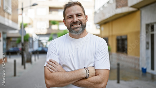 Smiling handsome hispanic man with grey hair and crossed arms standing outdoors in an urban city street.