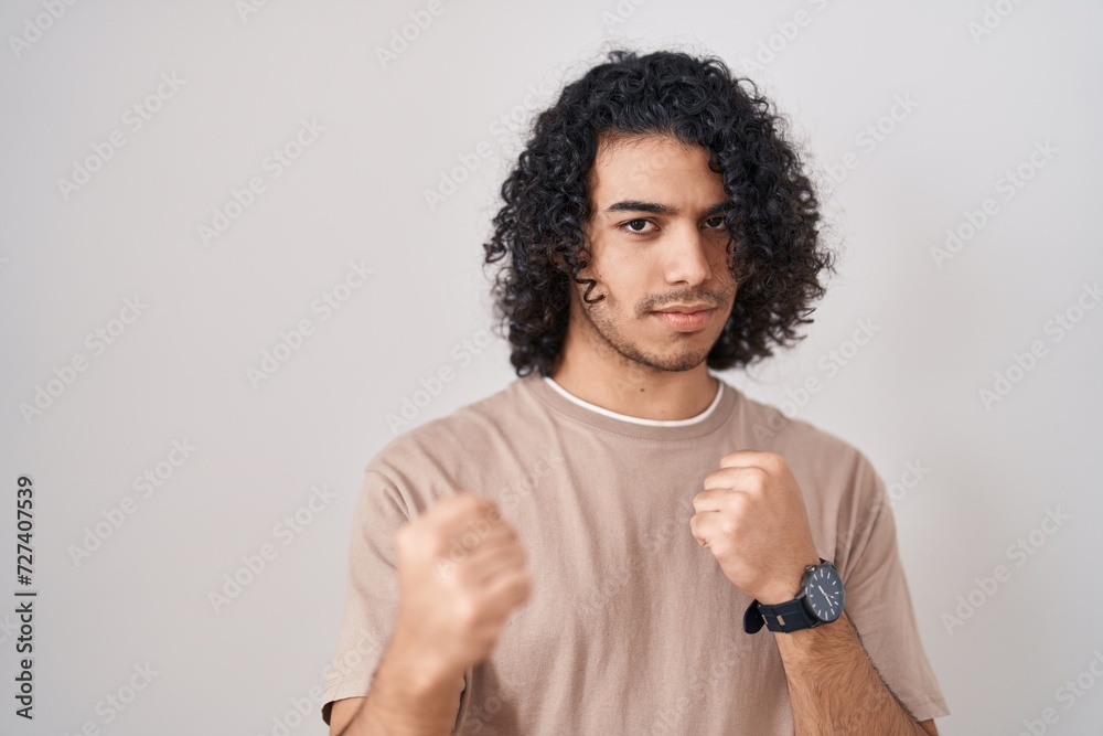 Hispanic man with curly hair standing over white background ready to fight with fist defense gesture, angry and upset face, afraid of problem Hispanic man with curly hair standing over white background ready to fight with fist defense gesture, angry and upset face, afraid of problem