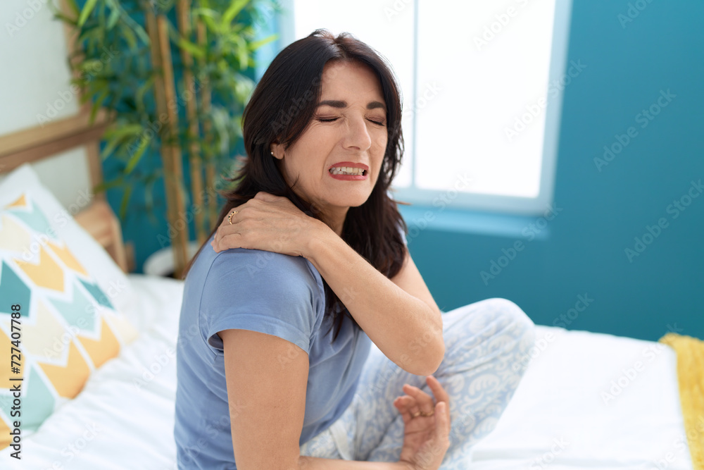 Middle age hispanic woman sitting on bed suffering for shoulder pain at bedroom Middle age hispanic woman sitting on bed suffering for shoulder pain at bedroom