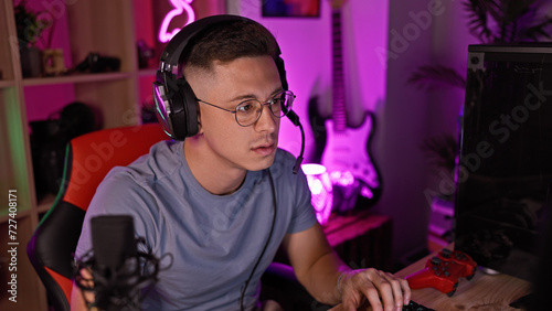 Hispanic man with headphones in a gaming room at night focused on a computer screen.