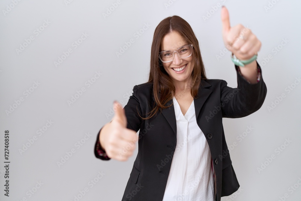 Beautiful brunette woman wearing business jacket and glasses approving doing positive gesture with hand, thumbs up smiling and happy for success. winner gesture. Beautiful brunette woman wearing business jacket and glasses approving doing positive gesture with hand, thumbs up smiling and happy for success. winner gesture.
