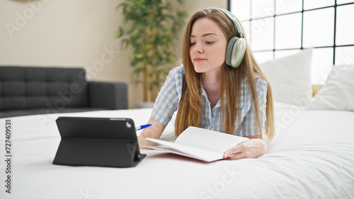 Young blonde woman student writing on notebook using touchpad studying on bed at bedroom