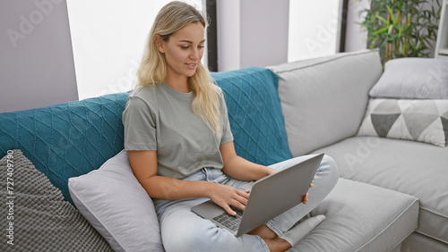 A young blonde woman relaxes at home, browsing on a laptop in her living room, exuding comfort and tranquility. © Krakenimages.com