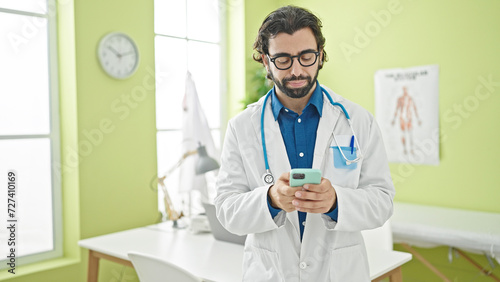 Young hispanic man doctor using smartphone thinking at the clinic
