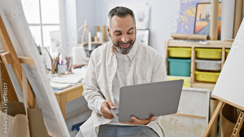 Smiling senior hispanic man with a beard using a laptop in a bright, artistic studio.