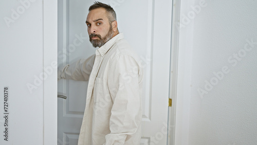 Mature bearded man in white attire opening a door, looking back with surprise at home entrance.