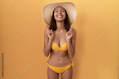 Young hispanic woman wearing bikini and summer hat gesturing finger crossed smiling with hope and eyes closed. luck and superstitious concept.