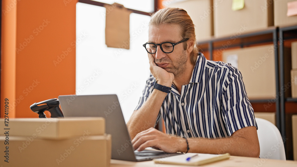 Focused man with glasses working on a laptop in a warehouse surrounded by cardboard boxes. Focused man with glasses working on a laptop in a warehouse surrounded by cardboard boxes.