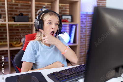Young caucasian woman playing video games wearing headphones surprised pointing with finger to the side, open mouth amazed expression.