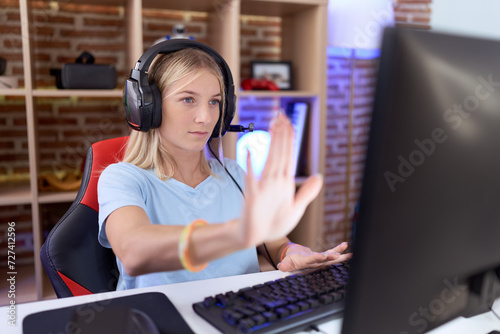 Young caucasian woman playing video games wearing headphones doing stop sing with palm of the hand. warning expression with negative and serious gesture on the face.