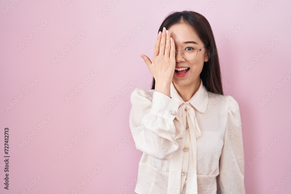 Young chinese woman standing over pink background covering one eye with hand, confident smile on face and surprise emotion. Young chinese woman standing over pink background covering one eye with hand, confident smile on face and surprise emotion.