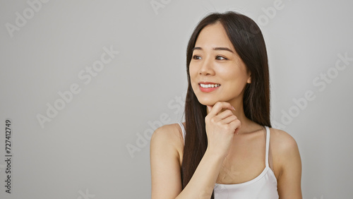 A thoughtful asian woman in a white tank top poses against an isolated white background, embodying casual elegance.