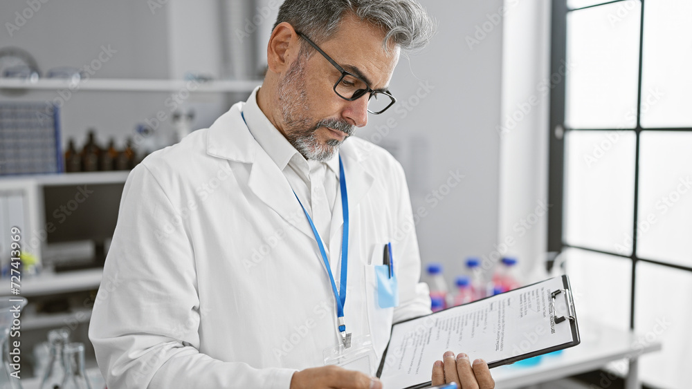 Handsome, grey-haired hispanic scientist engrossed in reading medical report on clipboard, working diligently in lab!