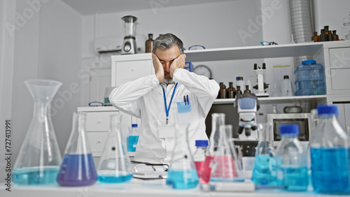 Stressed out grey-haired, young hispanic man, a professional scientist, under the microscope's spell, caught in overwork at an indoor laboratory