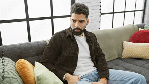 A contemplative man sits on a couch within a well-lit living room, exuding a casual yet stylish vibe.