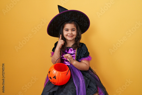 Adorable hispanic girl in witch costume, holds a pumpkin basket with a happy face, pointing one finger with an idea or question, isolated on a yellow background