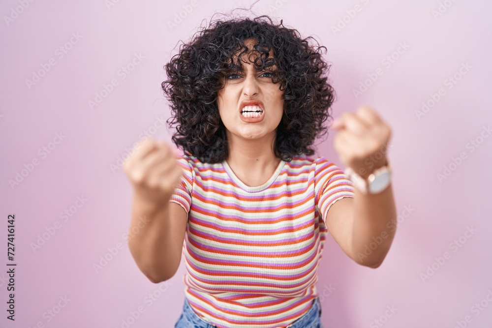 Young middle east woman standing over pink background angry and mad raising fists frustrated and furious while shouting with anger. rage and aggressive concept. Young middle east woman standing over pink background angry and mad raising fists frustrated and furious while shouting with anger. rage and aggressive concept.