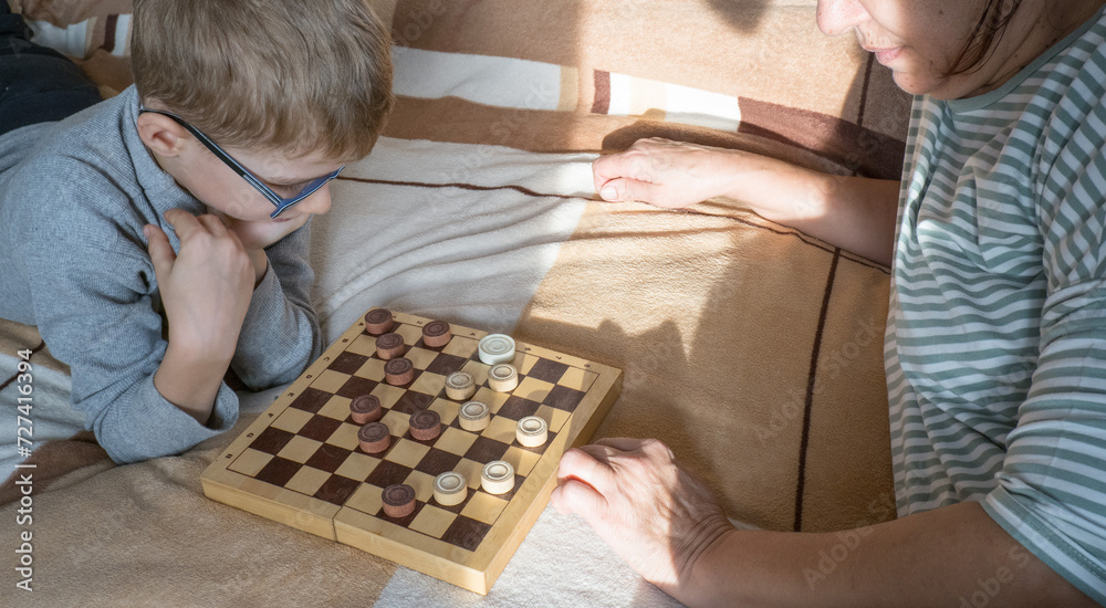 A caring middle-aged grandmother plays checkers with a child, a boy, lying on the bed. Happy family enjoying an interesting wooden board game at home.
