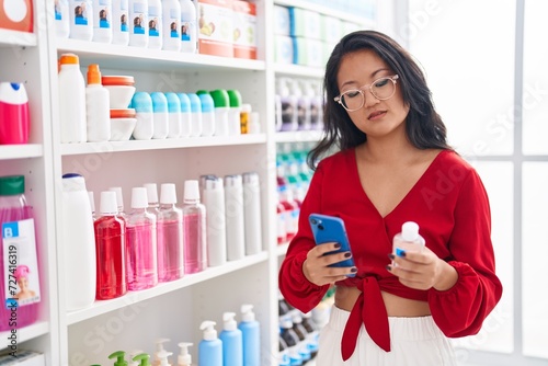 Young chinese woman customer using smartphone holding medicine bottle at pharmacy