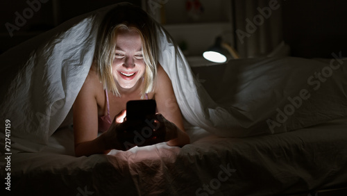 Young blonde woman using smartphone covering with blanket smiling at bedroom photo