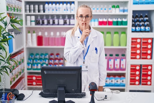 Young caucasian woman working at pharmacy drugstore asking to be quiet with finger on lips. silence and secret concept.