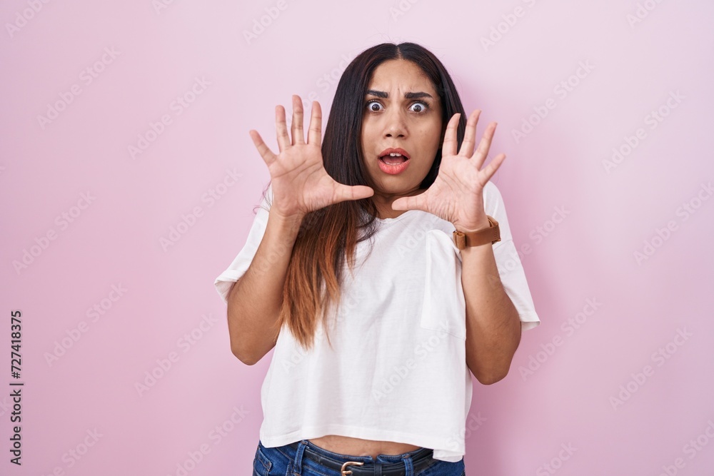 Young arab woman standing over pink background afraid and terrified with fear expression stop gesture with hands, shouting in shock. panic concept.
