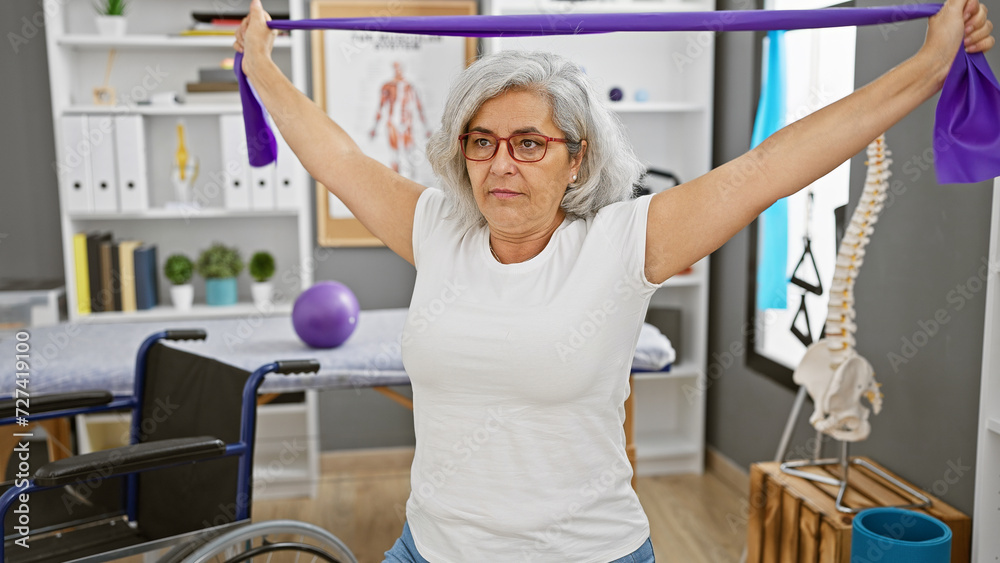 Mature woman exercising with resistance band in rehab clinic alongside wheelchair