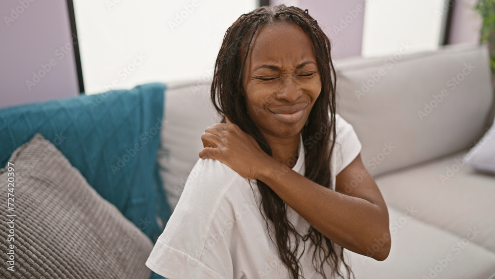 A young woman experiences shoulder pain in a modern apartment living room, grimacing while touching her aching joint. A young woman experiences shoulder pain in a modern apartment living room, grimacing while touching her aching joint.