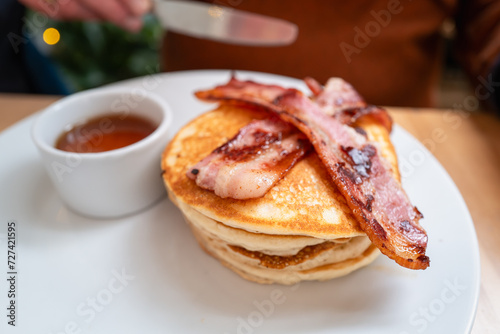 Two crispy bacon strips on a pile of pancakes. There is also a pot of golden maple syrup on the white plate.
